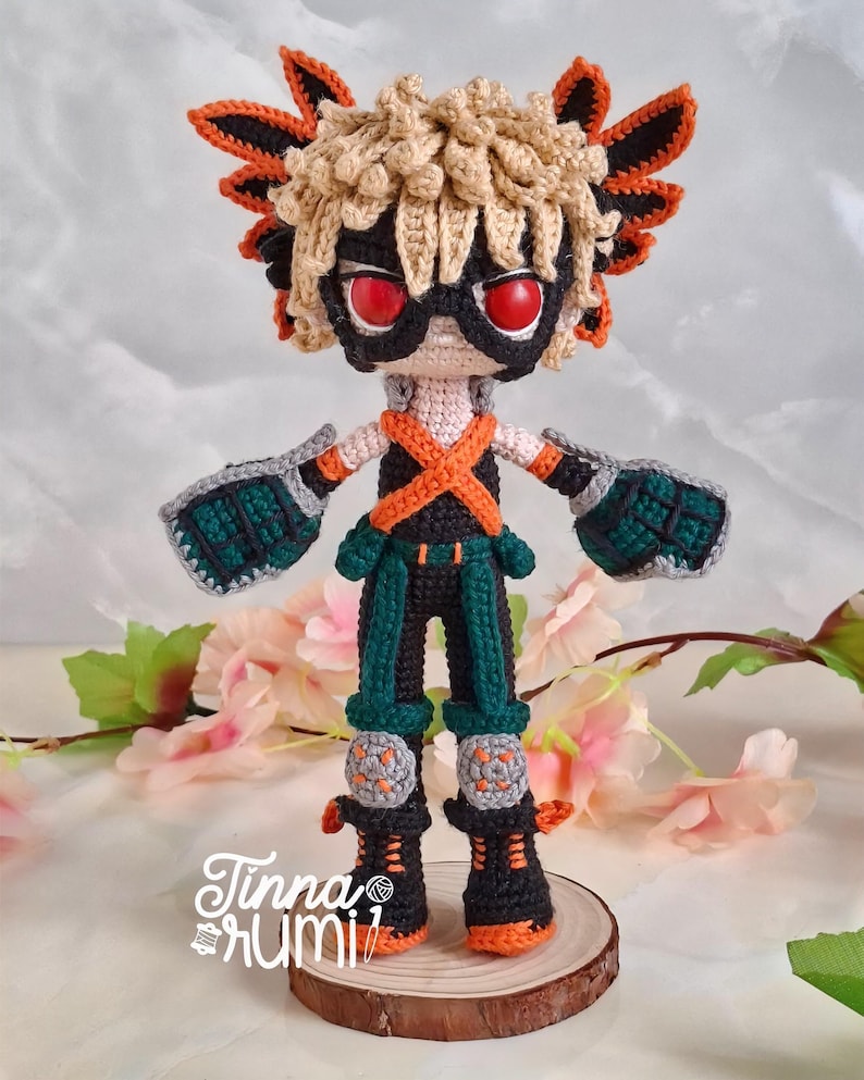 May include: A handmade crochet doll of a character with spiky blonde hair, red eyes, and a black mask. The doll wears a black and orange outfit with green accents, gray gloves, and knee pads. It stands on a wooden slice, with the artist's signature visible.