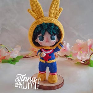 May include: A crochet doll featuring a character with dark green hair wearing a blue and yellow suit with a red chest emblem and a yellow hood with two tall, pointed ears. The doll stands on a circular wooden base with the text Tinna Rumi printed.