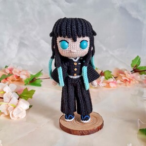 May include: Crocheted doll of a character with black hair, turquoise eyes, and a black outfit with gold buttons and turquoise accents. The doll is standing on a small wooden base. The background features pink and white flowers.