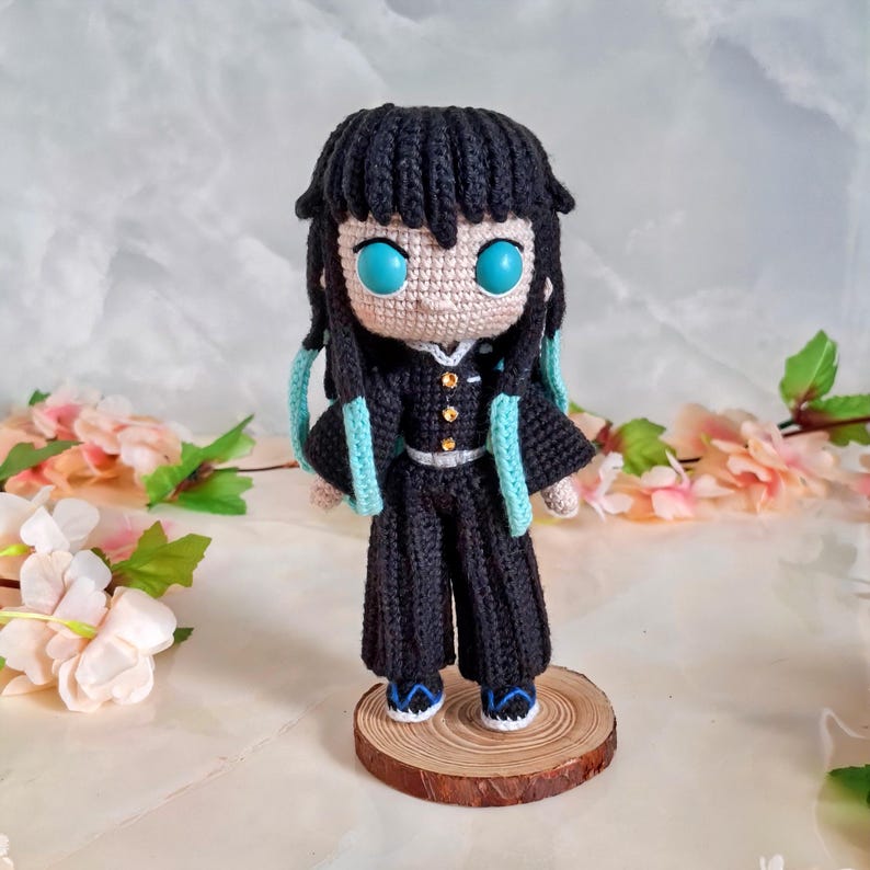May include: Crocheted doll of a character with black hair, turquoise eyes, and a black outfit with gold buttons and turquoise accents. The doll is standing on a small wooden base. The background features pink and white flowers.