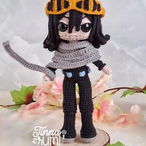 May include: A handmade crochet doll of a character with black hair, a gray scarf, and yellow goggles. The doll is wearing black pants with blue accents and is standing on a wooden base. The doll's face has large, expressive eyes. The doll is approximately 8 inches tall.