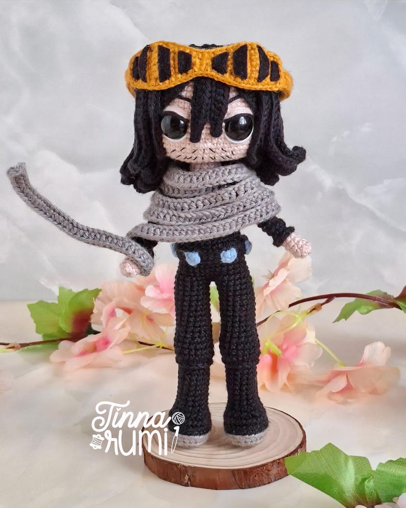 May include: A handmade crochet doll of a character with black hair, a gray scarf, and yellow goggles. The doll is wearing black pants with blue accents and is standing on a wooden base. The doll's face has large, expressive eyes. The doll is approximately 8 inches tall.