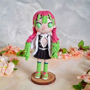 May include: A handmade crochet doll with pink and green hair, green eyes, and a black and white outfit. The doll is standing on a wooden base and is surrounded by pink flowers. The doll's hair is styled in two long braids.