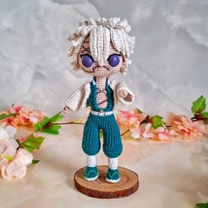 May include: Crocheted doll of a character with white hair, purple eyes, and facial scars. The doll wears a teal jumpsuit, a white jacket, and green and white shoes. It stands on a small wooden base, with a floral background.