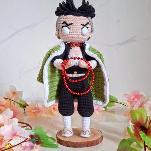 May include: Crocheted doll of a character with black hair, gray accents, and a stern expression. The doll wears a green and white cape, black pants, and a red beaded necklace. It stands on a wooden base, showcasing detailed craftsmanship.
