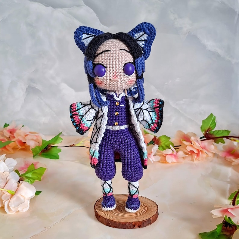 May include: A handmade crochet doll featuring a character with purple eyes, a purple jumpsuit, and a white jacket with butterfly wing patterns. The doll has blue hair and butterfly-shaped hair accessories. It stands on a small wooden base.