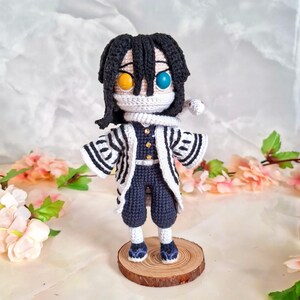 May include: A handmade crochet doll featuring a character with black hair, a white face covering, and contrasting yellow and blue eyes. The doll wears a dark blue outfit with a white and black striped jacket. It stands on a small wooden base.