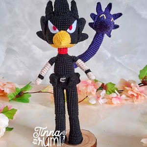 May include: A handmade crochet doll of a character with a black body, a yellow beak, and red eyes. The doll has a purple appendage and is mounted on a wooden base. The doll's arms have white and black details. The doll is signed "Tinna rumi".
