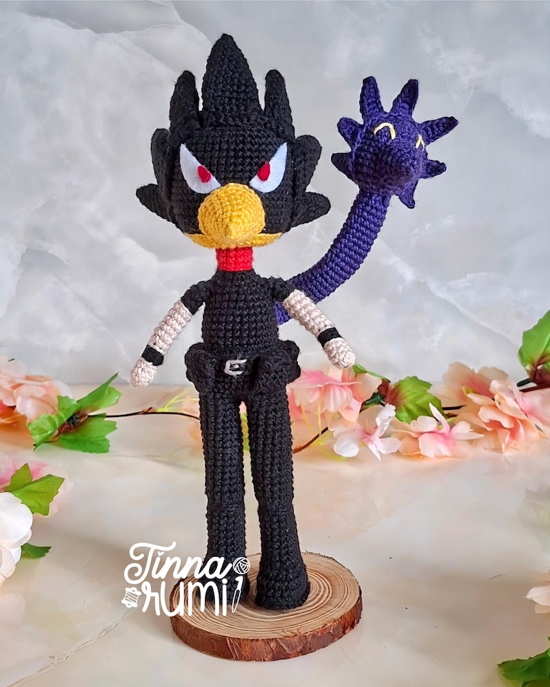 May include: A handmade crochet doll of a character with a black body, a yellow beak, and red eyes. The doll has a purple appendage and is mounted on a wooden base. The doll's arms have white and black details. The doll is signed "Tinna rumi".