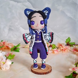 Puede incluir: Muñeca de ganchillo hecha a mano que representa un personaje con ojos morados, un mono morado y una chaqueta blanca con diseños de alas de mariposa. La muñeca tiene pelo azul y accesorios para el pelo con forma de mariposa. Se encuentra sobre una base de madera.
