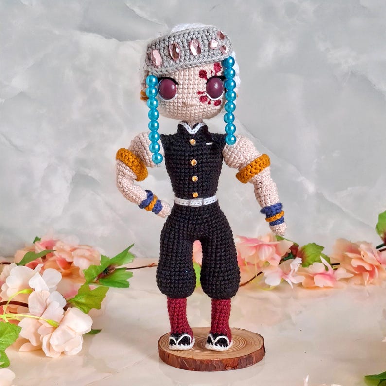 May include: A handmade crochet doll of a character with light skin, blue beaded hair, and a gray headband with pink gems. The doll wears a black outfit with gold and blue accents, and stands on a wooden base. The doll has red markings on its face.