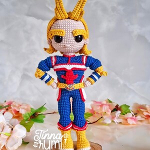 May include: A handmade crochet doll of a character in a blue, red, and yellow costume. The doll has yellow hair and is standing on a wooden base. The doll's costume has a red star on the chest. The doll is approximately 8 inches tall.