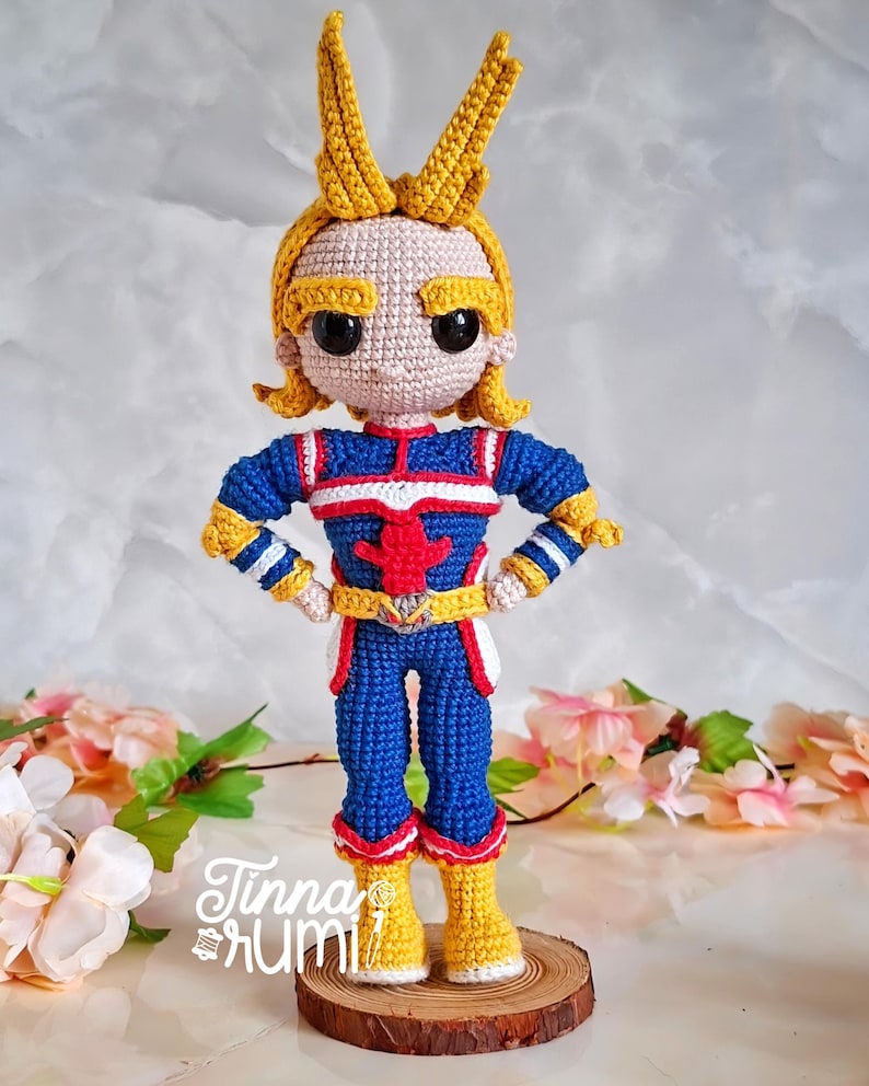 May include: A handmade crochet doll of a character in a blue, red, and yellow costume. The doll has yellow hair and is standing on a wooden base. The doll's costume has a red star on the chest. The doll is approximately 8 inches tall.