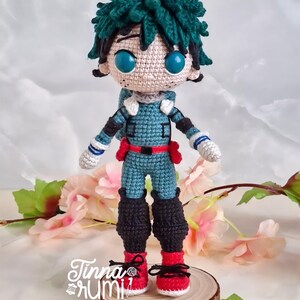 May include: A handmade crochet doll of a character with green hair and large blue eyes. The doll wears a teal and red outfit with black boots and red shoes. The doll is standing on a wooden slice, with the artist's signature in the bottom left corner.