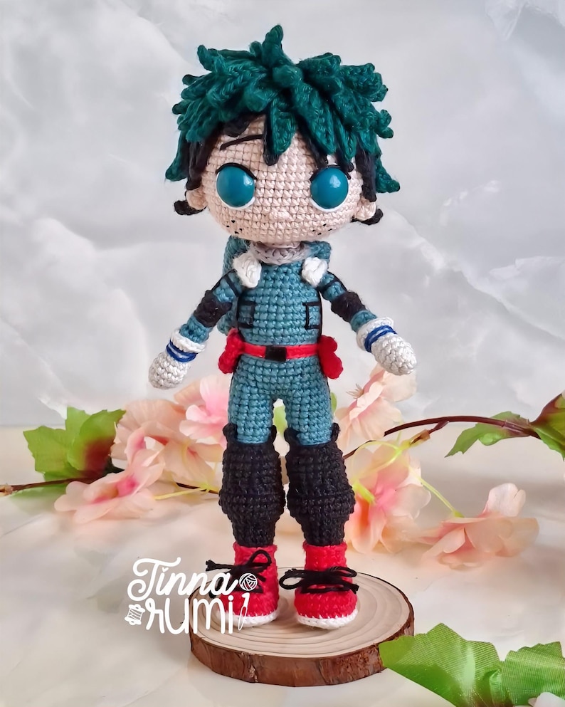 May include: A handmade crochet doll of a character with green hair and large blue eyes. The doll wears a teal and red outfit with black boots and red shoes. The doll is standing on a wooden slice, with the artist's signature in the bottom left corner.