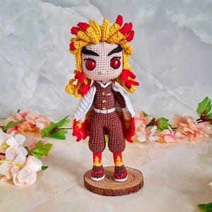 May include: A handmade crochet doll of a character with yellow and red hair, red eyes, and a brown outfit. The doll has a white cape and is standing on a wooden base. The doll's hair and outfit have flame-like details.