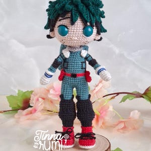 May include: A handmade crochet doll of a character with green hair and teal and red outfit. The doll has large blue eyes and is standing on a wooden slice. The doll's outfit includes a red belt and red shoes with black laces. The doll is a handmade item.