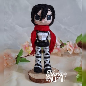 Puede incluir: Muñeca de ganchillo de un personaje con cabello negro, ojos grandes y una bufanda roja. La muñeca viste un top blanco, una falda negra y leggings plateados y negros. Está sobre un disco de madera. El texto "Tinna Rumi" es visible.