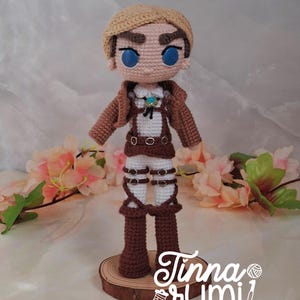 Puede incluir: Muñeca de ganchillo hecha a mano de un personaje con cabello castaño, ojos azules y una chaqueta marrón. La muñeca lleva una camisa blanca, pantalones marrones con hebillas plateadas y botas marrones. El texto "Tinna Rumi" es visible.