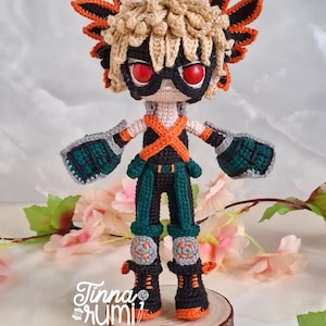 Puede incluir: Muñeca de ganchillo hecha a mano de un personaje con pelo rubio puntiagudo, ojos rojos y una máscara negra. La muñeca lleva un atuendo negro y naranja con detalles verdes y guantes grises. Se encuentra sobre una rodaja de madera.