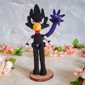 May include: A handmade crochet doll depicting a black bird-like character with a yellow beak, red eyes, and a purple arm. The doll is standing on a wooden base, with a red collar and white arm accents. The doll is approximately 20 cm tall.
