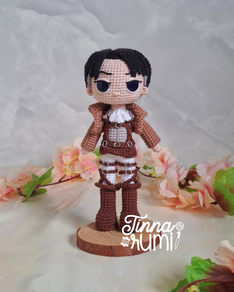 May include: Crocheted doll of a character with black hair, wearing a brown jacket, white shirt, and brown pants. The doll is standing on a small wooden base. The doll's eyes are blue. The doll is approximately 6 inches tall.