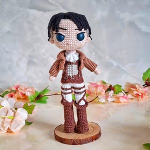 Puede incluir: Muñeca de ganchillo de un personaje con cabello negro y ojos azules, vestida con una chaqueta marrón, camisa blanca y pantalones marrones con correas. La muñeca está sobre una rodaja de madera, con un fondo floral.