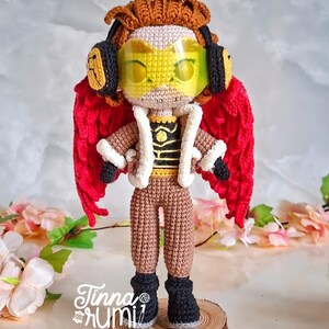 May include: A handmade crochet doll of a character with red wings, brown hair, and yellow goggles. The doll wears a brown outfit with a white-trimmed jacket and black boots. The doll is standing on a wooden base. The text "Tinna Rumi" is visible.