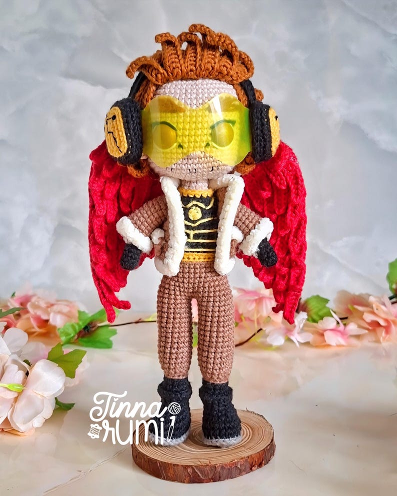 May include: A handmade crochet doll of a character with red wings, brown hair, and yellow goggles. The doll wears a brown outfit with a white-trimmed jacket and black boots. The doll is standing on a wooden base. The text "Tinna Rumi" is visible.