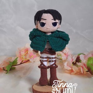 May include: A handmade crochet doll of a character with black hair, wearing a green cape, brown boots, and white pants. The doll is standing on a wooden base. The doll's eyes are blue. The doll is a character from the anime series Attack on Titan.
