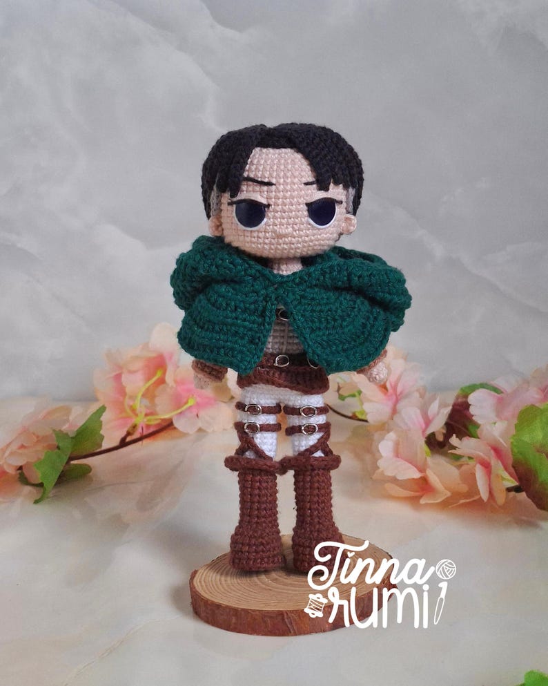 May include: A handmade crochet doll of a character with black hair, wearing a green cape, brown boots, and white pants. The doll is standing on a wooden base. The doll's eyes are blue. The doll is a character from the anime series Attack on Titan.