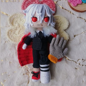 May include: A handmade crochet doll with white hair, red eyes, and a black suit with a red cape. The doll has a gray glove with an orange cuff and is wearing red sneakers. The doll is a unique, handcrafted item.