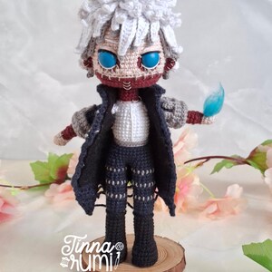 May include: A handmade crochet doll of a character with white hair, blue eyes, and a stitched-up face. The doll wears a black coat, pants, and a white shirt. It holds a blue flame. The doll is standing on a wooden base.