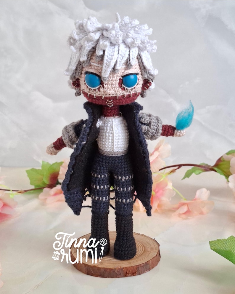 May include: A handmade crochet doll of a character with white hair, blue eyes, and a stitched-up face. The doll wears a black coat, pants, and a white shirt. It holds a blue flame. The doll is standing on a wooden base.