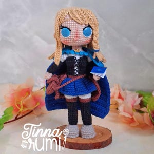 May include: Crocheted doll with blonde hair, blue eyes, and a blue cape. The doll wears a black and blue dress, holding a small blue book. A brown satchel hangs from her shoulder. The doll stands on a wooden base. The text "Tinna rumi" is visible.