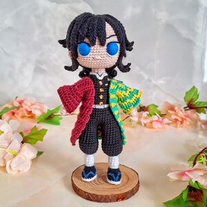 May include: A handmade crochet doll of a character with black hair and bright blue eyes. The doll wears a black outfit with a red and yellow patterned jacket. It stands on a small wooden base, showcasing the intricate details of the crochet work.
