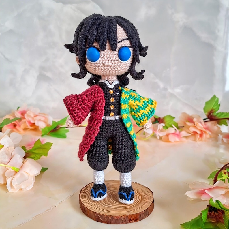 May include: A handmade crochet doll of a character with black hair and bright blue eyes. The doll wears a black outfit with a red and yellow patterned jacket. It stands on a small wooden base, showcasing the intricate details of the crochet work.
