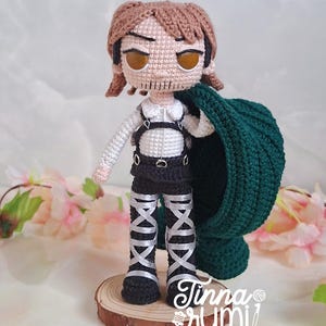 Puede incluir: Muñeca de ganchillo hecha a mano de un personaje con cabello castaño, camisa blanca, pantalones cortos negros y botas negras con detalles plateados. La muñeca sostiene una capa verde y está de pie sobre una base de madera. La cara de la muñeca tiene detalles cosidos.