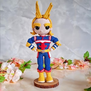May include: A crocheted doll depicting a superhero in a blue and red costume with yellow accents. The doll has yellow hair and boots, and is posed with hands on hips. It stands on a small wooden base. The doll's costume has a red emblem on the chest.