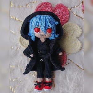 Puede incluir: Mu&ntilde;eca de ganchillo hecha a mano con cabello azul, ojos rojos y un atuendo negro con abrigo con capucha. La mu&ntilde;eca lleva zapatillas rojas y blancas. El fondo incluye corazones y una flor decorativos de ganchillo.