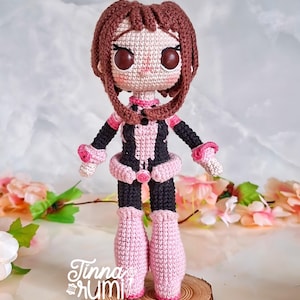 Może przedstawiać: Ręcznie robiona szydełkowa lalka postaci z brązowymi włosami i dużymi brązowymi oczami. Lalka ma na sobie czarno-różowy strój z pasującymi butami i mankietami na nadgarstki. Stoi na małym kawałku drewna, z imieniem artysty.