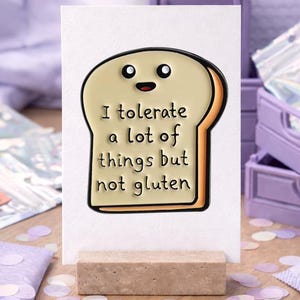 Könnte beinhalten: Eine Grußkarte mit einem Cartoon-Brotstück mit einem lächelnden Gesicht. Auf der Karte steht "Ich toleriere viele Dinge, aber kein Gluten." Sie wird auf einem kleinen, rechteckigen Steinsockel präsentiert. Der Hintergrund umfasst lila Kisten und Konfetti.
