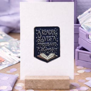 Può includere: Una spilla in smalto blu scuro a forma di scudo, con l'illustrazione di un libro aperto e il testo A READER LIVES A THOUSAND LIVES in lettere argentate. La spilla è fissata su un cartoncino rettangolare bianco poggiato su un blocco di legno.