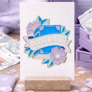 Könnte beinhalten: Eine weiße Karte mit einem "BOOK LOVER" Anstecker, der ein blaues Buchdesign mit Blumen und einem Banner zeigt. Der Anstecker wird auf einem Holzständer präsentiert, umgeben von lila Konfetti und kleinen lila Boxen.