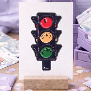 Könnte beinhalten: Ein schwarzer Ampel-Anstecker mit roten, gelben und grünen Lichtern und den Worten „DON'T KNOW“. Der Anstecker wird auf einer weißen Karte auf einem Holzständer mit einem Hintergrund aus lila Kisten und Konfetti präsentiert.