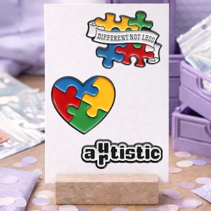 Könnte beinhalten: Eine weiße Karte mit einem herzförmigen Puzzleteil-Design und dem Wort "artistic" darunter. Ein Banner über dem Herzen lautet "DIFFERENT NOT LESS". Die Puzzleteile sind rot, gelb, blau und grün.