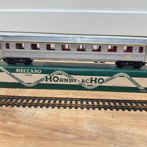 May include: A silver model train car with red interior details sits atop a green and white box labeled "MECCANO PARIS HORNBY-ACHO". A section of train tracks is in front of the box. The train car is a vintage collectible.
