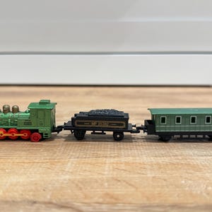 Puede incluir: Un juego de tren de juguete en miniatura con una locomotora verde con ruedas rojas, un vagón de carbón negro y un vagón de pasajeros verde. El tren está sobre una superficie de madera.