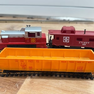 Puede incluir: Un juego de trenes modelo con una locomotora roja y plateada, un vagón de cola rojo y un vagón de carga naranja. El vagón de carga tiene el texto "Union Pacific" y "Main Tence of Way". El juego está sobre una superficie de madera.