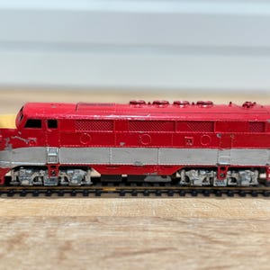 Puede incluir: Una locomotora de tren de juguete roja y plateada vintage con un frente amarillo. El tren modelo está sobre un tramo de vía, mostrando su diseño clásico. El tren es un artículo de colección.